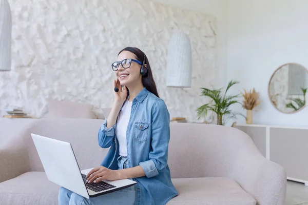 Online work. Young beautiful freelancer woman in glasses and jeans sitting at home on sofa and working with laptop and headphones online, talking on video call with clients, partners.
