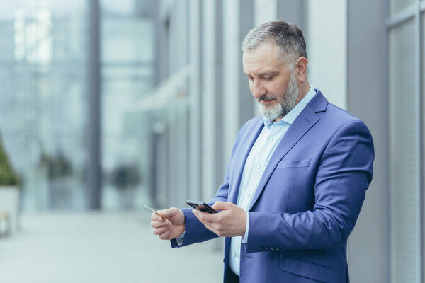 A serious and focused senior male businessman is standing near an office center, holding a mobile phone and a credit card in his hands, entering the card number into the account on the phone.