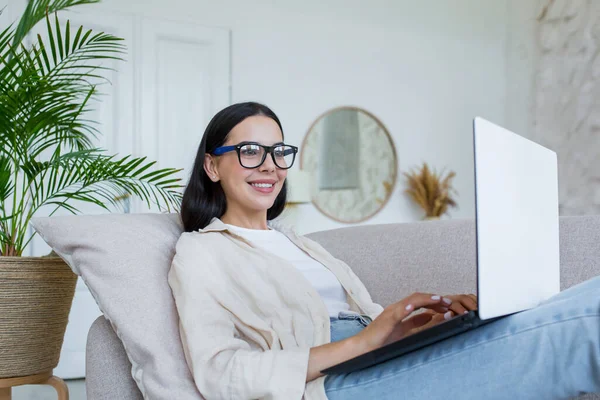 A young beautiful female writer in glasses sits comfortably on the sofa at home, holding a laptop on her lap. Type the text, print the book, smile.