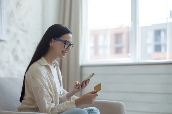 Online shopping. Satisfied young beautiful woman in glasses sitting on sofa near window at home. Holds a credit card and phone, pays for the product, orders delivery.