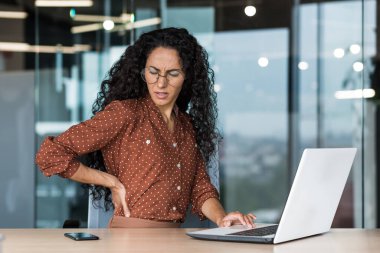 Ofis çalışanı Latin Amerikalı iş adamı iş yerinde hasta, aşırı yorgun bir iş kadını sırt ağrısı çekiyor, elleriyle sırtına masaj yapıyor..