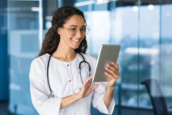 Latin American doctor woman using tablet computer, female doctor working inside office building wearing white medical coat.