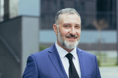 Gri saçlı, kahverengi saçlı ve kravatlı kıdemli yakışıklı işadamının portresi. Ofis merkezine yakın, kameraya bakıyor, gülümsüyor..