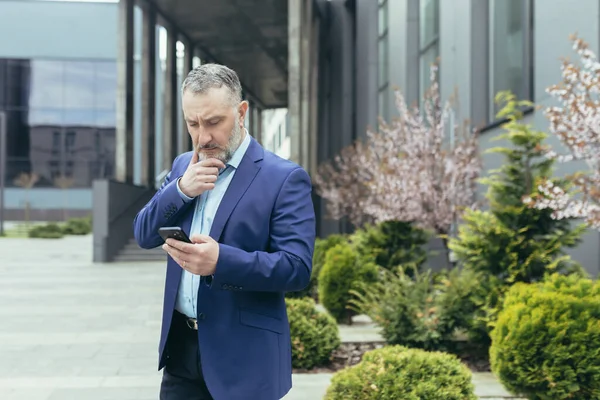 Pensive and worried senior man in a business suit stands on the street near a modern building, looks at a mobile phone, reads, types, holds his beard thoughtfully.