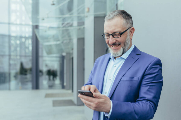 Happy senior handsome gray-haired man, businessman in suit and glasses. Holds the phone in his hands, received good news, reads the message. It stands on the street near a modern building