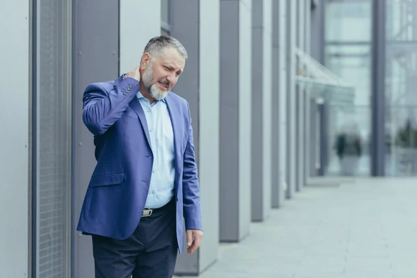 An senior man, a banker, an office worker in suit, went outside the office for a lunch break, his neck hurts, he massages his neck with one hand, he feels pain.