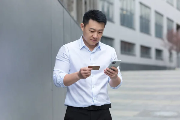 Portrait of a young asian man standing on the street near a modern office center, holding a phone and a credit card in his hands, entering the card number, making online purchases, placing an order.