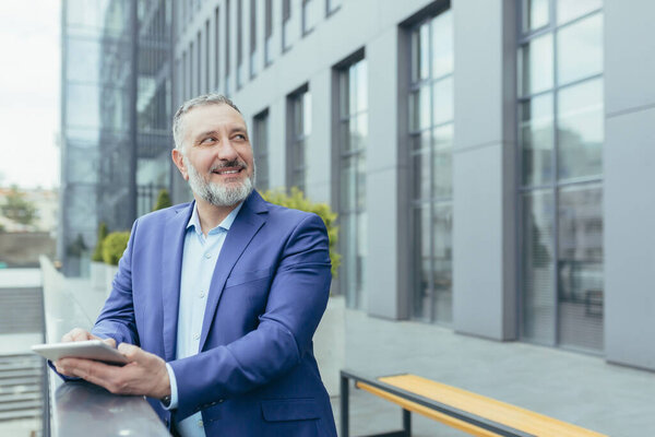 Senior handsome gray-haired male teacher, professor, lecturer is standing in a suit on the balcony in a modern campus, holding a tablet in his hands. He looks up at someone, smiles, talks.