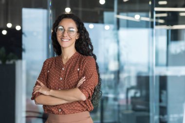 Genç, güzel ve başarılı İspanyol iş kadını gülümseyen ve kameraya bakan, ofis içinde çalışan, gözlüklü, kıvırcık saçlı, kollarını kavuşturmuş portre çizen bir kadın.