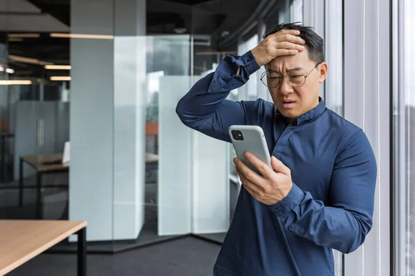 Asian businessman inside office building upset and sad reading bad news on mobile phone, business owner investor using smartphone standing near window