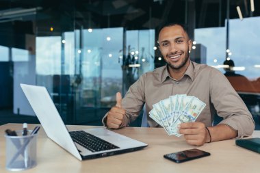 Başarılı genç yatırımcı portresi, gülümseyen ve kameraya bakan adam, modern ofis binasında çalışan, elinde dolar parası tutan ve olumlu başparmak gösteren.