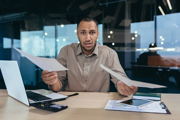 Portrait of disappointed young hispanic financier, businessman looking at camera in despair and holding failed financial report of business in hands, man working inside office building paperwork.