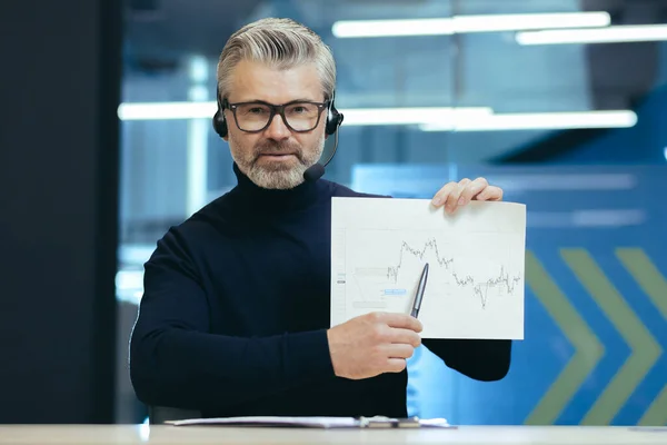 Senior teacher business teacher coach teaches online finance, gray-haired man with headset and glasses looks into camera explains financial chart to interlocutor, video call online learning.