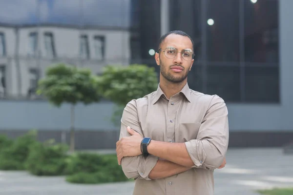 Serious and pensive young office worker with crossed arms looking at camera, businessman in shirt outside office building pensive.