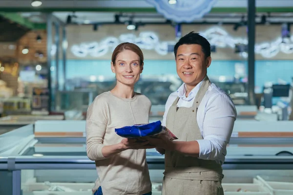 Asian male seller in supermarket recommending product to female buyer, couple looking at camera and smiling satisfied with shopping service near refrigerators in store.