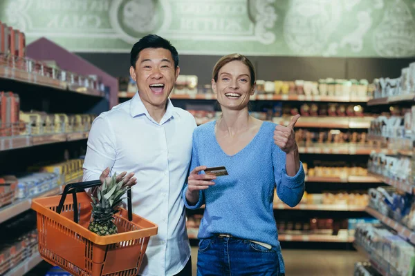 Happy diverse family couple man and woman shoppers in supermarket, looking at camera and smiling happy shopping, holding phone and bank credit card in hand, with shopping basket.