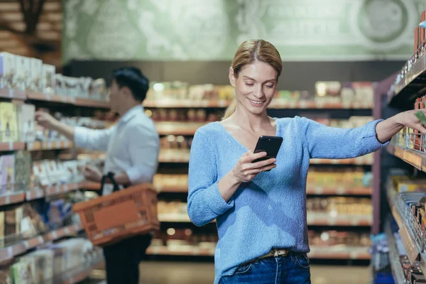 Young beautiful woman buyer in a supermarket chooses goods in the store, housewife looks at the phone, uses an application for selecting products.