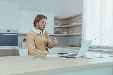 Evden uzakta çalışan genç bir işadamı mutfakta oturuyor, bilgisayarını video için kullanıyor konsantre oluyor ve iş arkadaşlarıyla internette ciddi bir görüşme yapıyor..