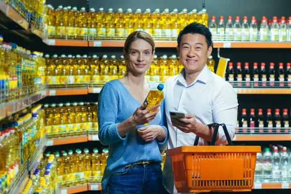 A young international couple, a woman and an Asian man, are choosing oil in a supermarket. They are holding a bottle of oil, a basket and a phone. They are looking at the camera and smiling.