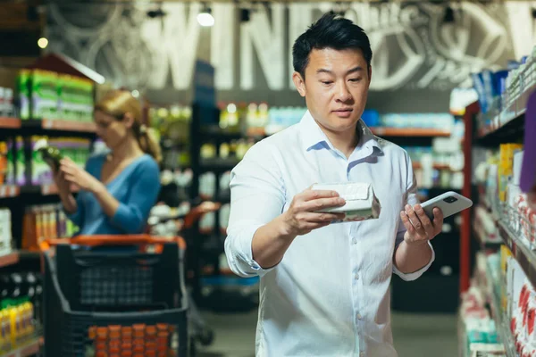 A young Asian man is standing in a supermarket, choosing pads for his wife, girlfriend. He does not know which one to choose, he stands thoughtfully, confused, holding the phone in his hand.