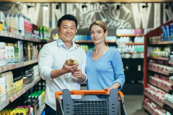 A young international couple, a woman and an Asian man are walking between the rows with a trolley in the supermarket. The man is holding a bottle of shampoo in his hands, looking at the camera