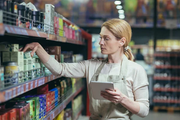 A beautiful young female employee of a supermarket, store in a work uniform displays goods on the shelf, counts, writes in a notebook.