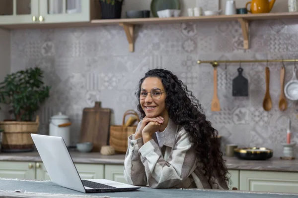 Portrait of young beautiful hispanic woman at home, female freelancer working remotely using laptop, looking at camera and smiling in glasses and curly hair in kitchen
