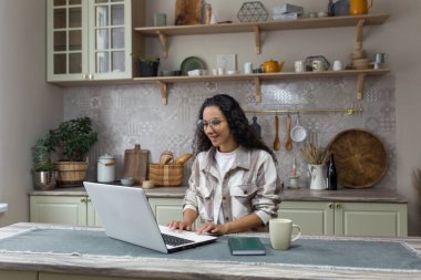 Evde internetten öğrenme, kıvırcık saçlı ve gözlüklü genç Latin Amerikalı kadın dizüstü bilgisayar kullanıyor, mutfakta oturan kız öğrenci....