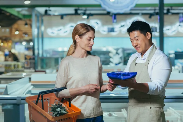 Consultant in a supermarket. A young beautiful woman wants to buy frozen products in the store. An employee consultant, an Asian man, helps her choose. They talk, smile