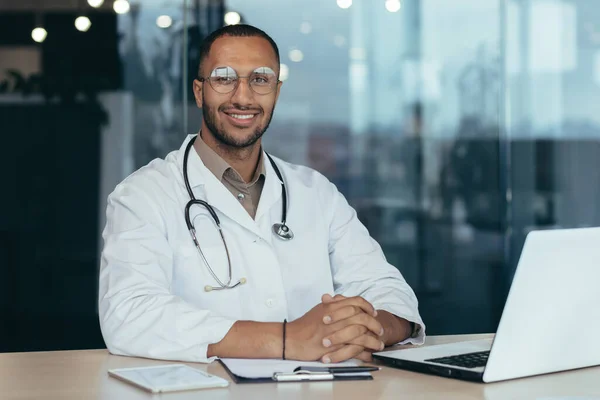 African american doctor portrait, man working inside modern clinic office at table using laptop, doctor in medical coat and stethoscope smiling and looking at camera.