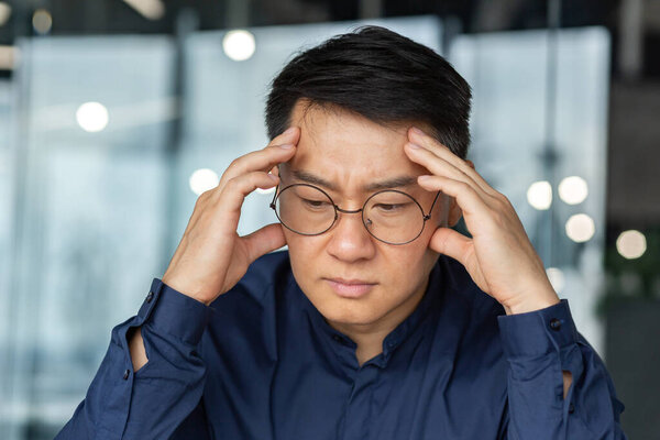 Closeup thinking businessman, Asian man in glasses and shirt holding hands on head making important decision, male manager working inside modern office building, thoughtful and serious boss.