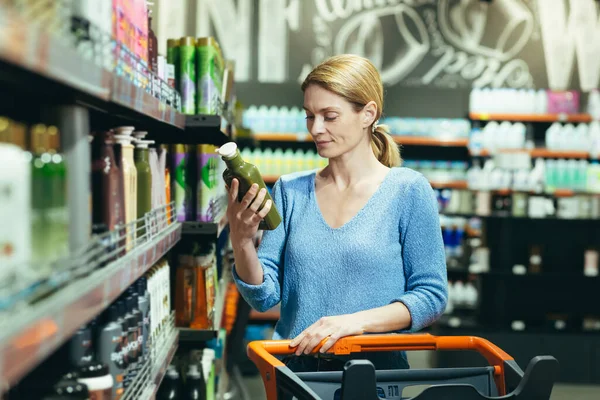 A young woman stands in a store in the department of household chemicals with a cart for goods, chooses shampoo for hair, holds a green one in her hand, reads the composition of the gel, shampoo.
