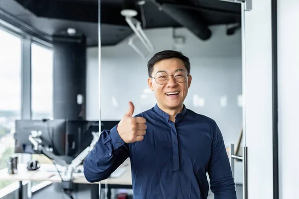 Portrait of successful asian businessman, man working in modern office looking at camera and smiling, broker trader showing thumbs up.