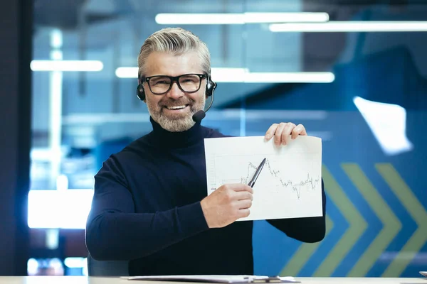 A man in headphones with a microphone. Sitting in the office, holding graphics, a project in his hands and pointing to the camera with a pen. Conducts business meeting, web conference, smiles.