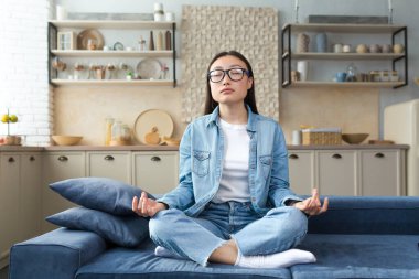 Beautiful Asian woman relaxing at home sitting on sofa and meditating in lotus pose, doing breathing exercises in living room.