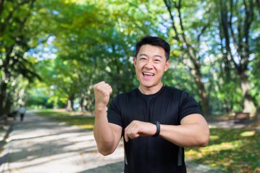 Mutlu ve başarılı bir adam kameraya bakıyor ve gülümsüyor, Asyalı sporcu antrenman sonuçlarından memnun, spor bilekliği takıyor, zaferi kutluyor, başarı jestini yapıyor..