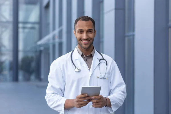 Portrait of happy and smiling hispanic doctor, man smiling and looking at camera, doctor using tablet computer for online consultation, outside of modern clinic.