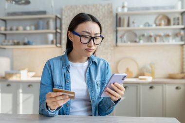 Çevrimiçi bir mağazadan alışveriş yapmaya çalışan Asyalı bir kadın, mutfakta bir kanepede oturan bir kadın akıllı telefonuyla başvuru yapıyor ve elinde bir banka kartı tutuyor..