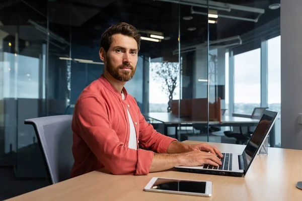 Portrait of mature programmer team leader man with beard and in red shirt is concentrating and thinking looking at camera , developer working inside modern company using laptop for coding.