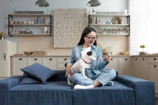 Young beautiful asian woman with pet jack russell terrier talking on video call with friends, woman at home in kitchen using smartphone for remote communication
