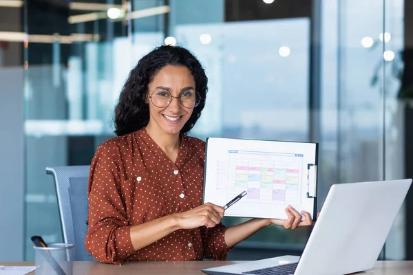 Portrait Hispanic business coach, woman smiling and looking at camera, showing documents with economic indicators to camera, teacher teaching remotely, mentor using laptop for online learning.