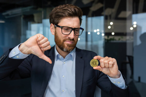 Close-up photo of male businessman holding gold crypto currency bitcoin coin in hands, mature investor disappointed with money rate and investment, boss in business suit showing finger down