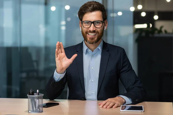 Video call on online meeting with colleagues, mature boss in business suit looking at web camera and smiling waving to employees, businessman in glasses working inside modern office building.