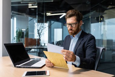 Modern ofis binasında dizüstü bilgisayarla çalışan ciddi ve odaklanmış olgun bir patron iş adamı bankadan gelen mektubu dikkatle okuyor gözlüklü ve sakallı üzgün bir adam..