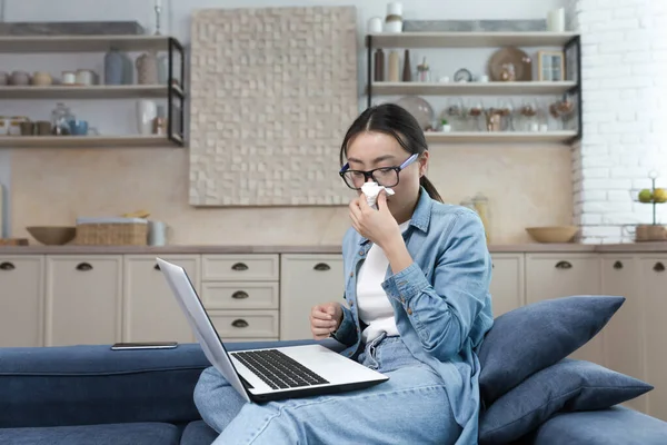 Work from home. Hospital. A young Asian woman freelancer fell ill. Works from home on the sofa at the laptop. He feels bad. He has a runny nose, wipes his nose with a napkin.