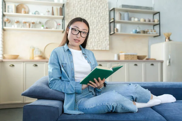 Portrait of a young beautiful Asian woman in denim clothes and glasses sitting at home on the sofa on the background of the kitchen. He holds a green book in his hands, reads, looks at the camera.