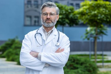 Ciddi ve düşünceli gri saçlı doktor kameraya bakıyor modern kliniğin dışında steteskoplu ve gözlüklü olgun bir adam..