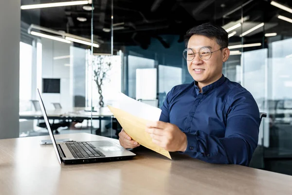 Happy asian man in glasses received letter opens and reads message document, businessman works inside modern office building on laptop, good news in notification.