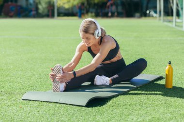 Sarışın kadın spor minderinde oturuyor, sporcu kadın kaslarını esnetiyor, kulaklıkla müzik dinliyor ve gülümsüyor, güneşli bir günde sabah stadyumunda..