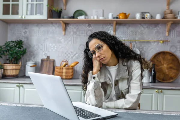 Sad and thinking woman working at home using laptop, Hispanic woman in kitchen at table wearing glasses and curly hair, businesswoman working remotely at home alone.